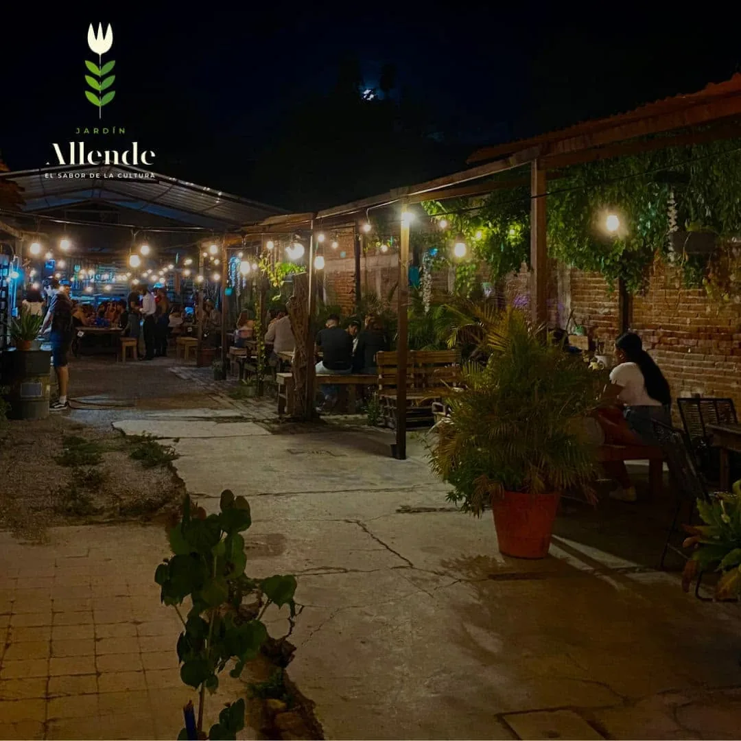 Jardín Allende iluminado por la noche con visitantes disfrutando de las mesas